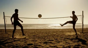 Fotografia profissional retratando a origem do futevôlei na Copacabana dos anos 60, com dois jogadores em silhueta sobre a areia, jogando bola sobre uma rede de vôlei improvisada contra um sol dramático de fim de tarde.