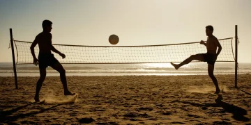 Fotografia profissional retratando a origem do futevôlei na Copacabana dos anos 60, com dois jogadores em silhueta sobre a areia, jogando bola sobre uma rede de vôlei improvisada contra um sol dramático de fim de tarde.
