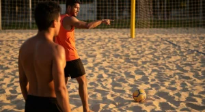 Dupla de futevôlei em quadra de areia ao entardecer, com um jogador gesticulando e o outro observando atentamente, ilustrando a comunicação e estratégia no jogo.