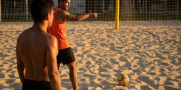 Dupla de futevôlei em quadra de areia ao entardecer, com um jogador gesticulando e o outro observando atentamente, ilustrando a comunicação e estratégia no jogo.