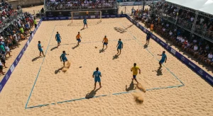 Cena aérea de um torneio internacional de futevôlei na praia, com atletas globais em quadra e uma multidão vibrante observando sob o sol forte, destacando a ascensão mundial do esporte.