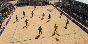 Cena aérea de um torneio internacional de futevôlei na praia, com atletas globais em quadra e uma multidão vibrante observando sob o sol forte, destacando a ascensão mundial do esporte.