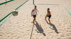 Futevôlei: Desvendando os Segredos dos Craques nas Areias Brasileiras Fotografia aérea de alta performance mostrando jogadores de futevôlei em uma quadra de areia vibrante, com o sol do meio-dia destacando a estratégia de posicionamento e o movimento tático durante um jogo profissional.