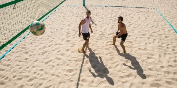 Fotografia aérea de alta performance mostrando jogadores de futevôlei em uma quadra de areia vibrante, com o sol do meio-dia destacando a estratégia de posicionamento e o movimento tático durante um jogo profissional.