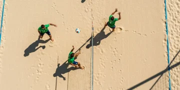 Vista aérea de drone capturando dois jogadores de futevôlei em quadra de areia, demonstrando posicionamento dinâmico e cobertura estratégica durante uma jogada, com padrões geométricos de sombra.