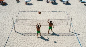 Vista aérea de uma partida de futevôlei na praia de Copacabana nos anos 60, com dois jogadores em ação, rede improvisada e areia quente.