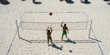 Vista aérea de uma partida de futevôlei na praia de Copacabana nos anos 60, com dois jogadores em ação, rede improvisada e areia quente.