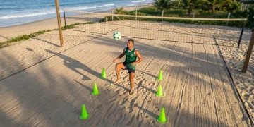 Fotografia aérea de um jogador de futevôlei, usando camisa verde, realizando um treino tático com cones verdes em uma quadra de areia. O sol da manhã ilumina a cena, enfatizando a precisão e o foco do atleta.