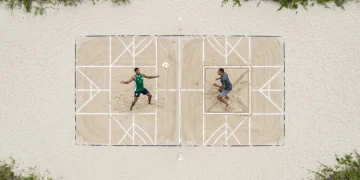 Vista aérea de drone de uma quadra de futevôlei na areia, com dois atletas em um treino combinado de fundamentos clássicos e inovações táticas, com marcações geométricas no chão, sob luz natural difusa.