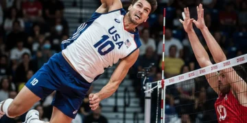 Male volleyball player powerfully spiking ball over net in national team jersey.