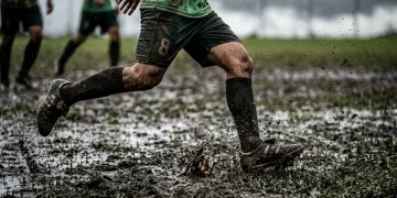 Fotografia documental de alto contraste de chuteiras de futebol sujas de lama, mostrando a determinação tática de um jogador em campo molhado, com foco na disciplina e contra-ataques.