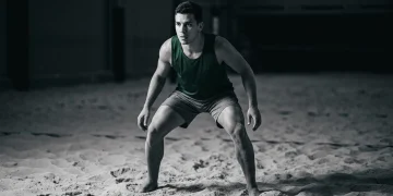 Atleta de futevôlei em quadra de areia escura, em postura defensiva, com iluminação dramática destacando a tensão nos quadris e ombros, focando na leitura de jogo. O atleta veste um top atlético verde e shorts de areia.